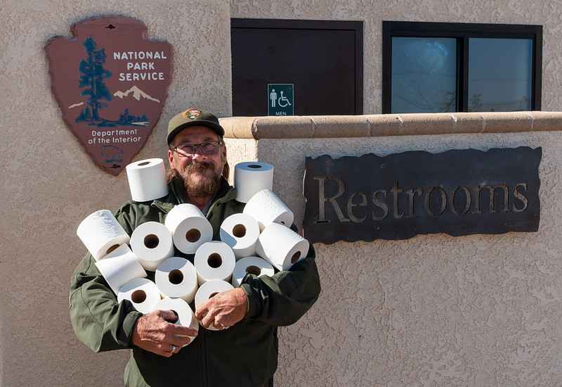 Ranger with a beard with arms full of rolls of toilet paper in front of a restroom near the entrance to Joshua Tree National Park
